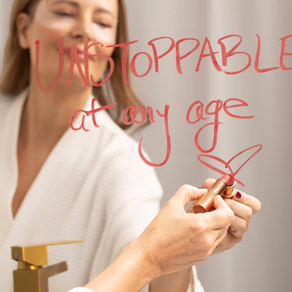 SIO Beauty woman in robe writing "UNSTOPPABLE at any age" on mirror with lipstick, applying Eye Hydroseal™ Patches.