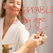 SIO Beauty woman in robe writing "UNSTOPPABLE at any age" on mirror with lipstick, applying Eye Hydroseal™ Patches.